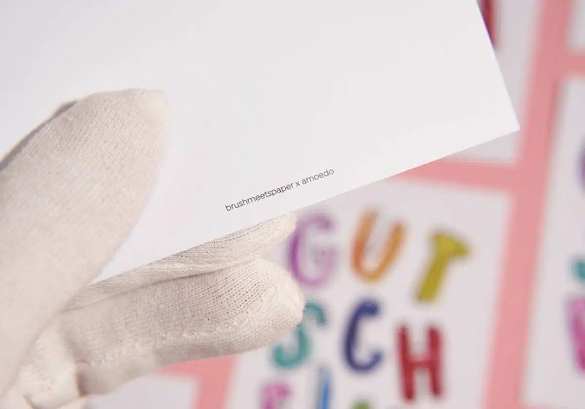 Eine behandschuhte Hand hält eine weiße Karte von brushmeetspaper x amoedo mit einem kleinen aufgedruckten Text. Im Hintergrund sind bunte Buchstaben zu sehen, leicht verschwommen, als würden sie „GUTSCHEIN“ auf einer weiteren farbenfrohen Postkarte »GUTSCHEIN« buchstabieren.