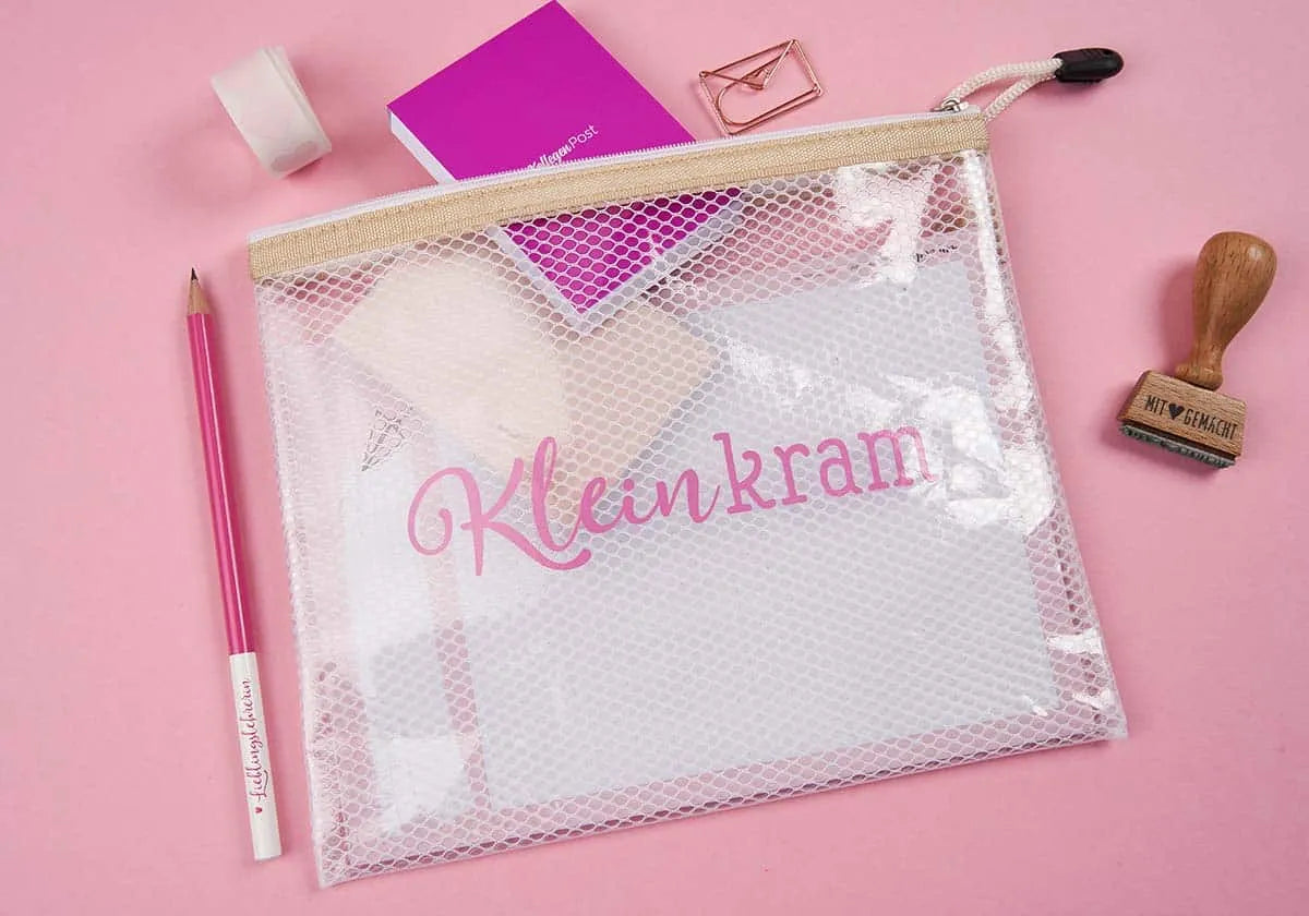 Ein PVC-freies Netz-Mäppchen mit der Aufschrift „Kleinkram“ von amoedo mit Reißverschluss ordnet Schreibwaren auf einer rosa Oberfläche. Daneben befinden sich ein rosa Bleistift, Klebeband, ein lila Notizblock und ein Stempel.