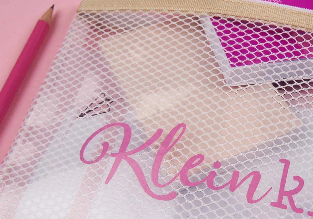 Ein rosa Bleistift liegt neben einem transparenten Mäppchen »Kleinkram« von amoedo mit Reißverschluss aus Mesh. Dieses Mäppchen mit seiner strapazierfähigen Oberfläche bietet Platz für verschiedene Kosmetika und trägt auf der Vorderseite das Wort „Kleink“ in rosa Schreibschrift auf einem zartrosa Hintergrund.